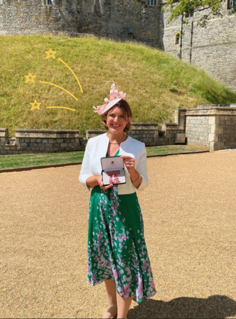 A picture of Action Tutoring's former CEO Susannah Hardyman, displaying her MBE award in front of Windsor Castle. Susannah is smiling at the camera and holding the award in front of her and wearing a floral hat, jacket and dress.