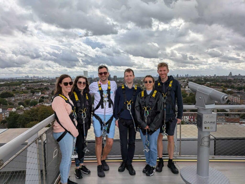 A picture of the abseil fundraising team at the top of the Tottenham Hotspur Stadium. They are wearing safety gear and are sitting beside grey railings in front of a cloudy London skyline. 