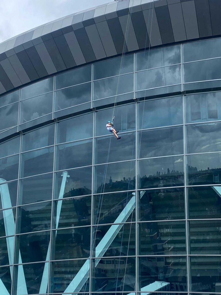 A picture of fundraiser Adam taking on the free-fall abseil for Action Tutoring aside the Tottenham Hotspur Stadium. Adam is holding on to a safety rope while scaling the glass windows of the stadum. 