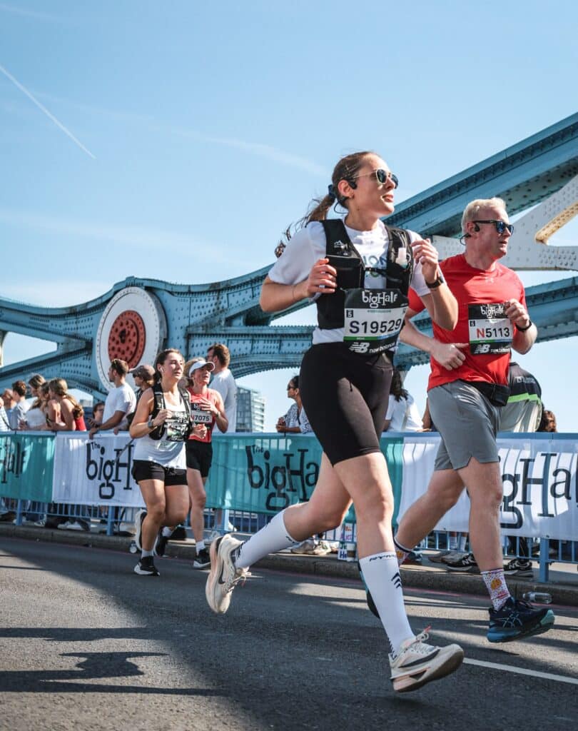 A picture of Kalina from QBE's Data Engineer and fundraiser running the Big Half for Action Tutoring. Kalina is wearing a white Action Tutoring t-shirt and black running vest and dark glasses and is running across Tower Bridge with three other participants and beside a crowd cheering them on. 