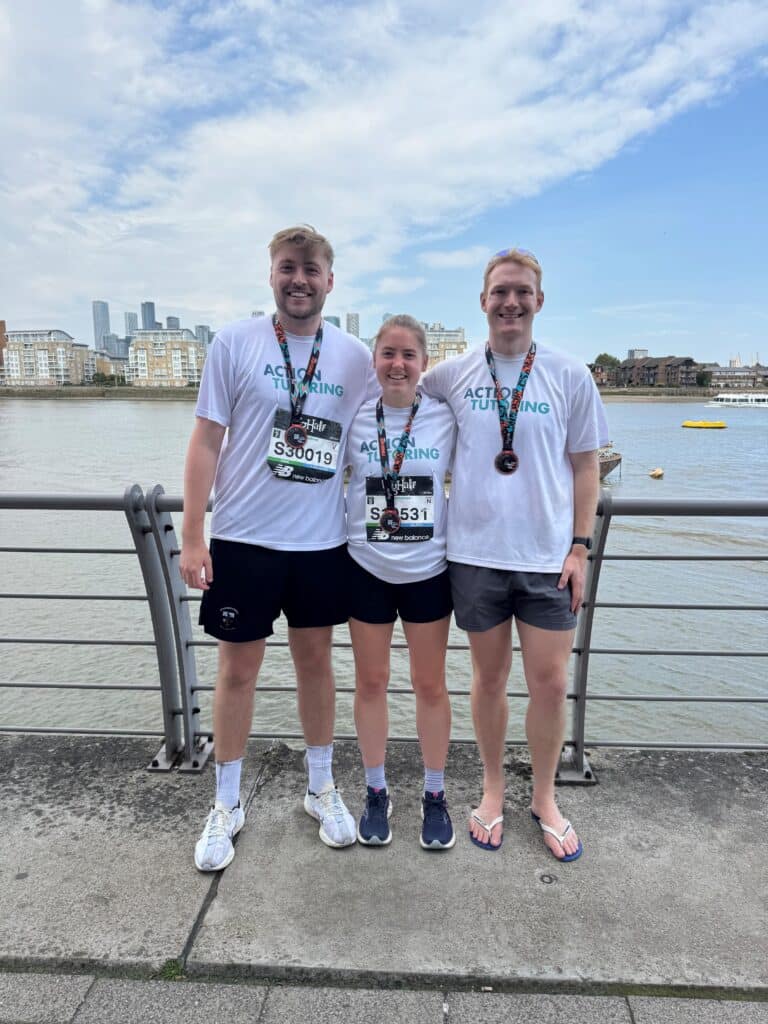 A picture of QBE fundraisers Freya, Patrick and Cameron standing in front the River Thames after completing the Big Half and posing with their medals. They are all smiling at the camera and wearing a white Action Tutoring t-shirt and shorts. 