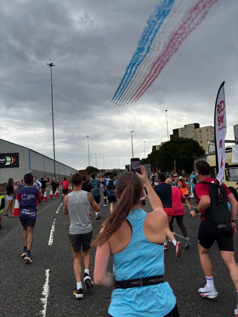 A picture of Action Tutoring's Corporate Partnerships Manager, Mollie, taking part in the Great North Run. She is running with many other participants and holding out her phone to take a picture of the RAF's Red Arrows who are completing an aerial display above with blue, white and red dye lining the grey skyline. 