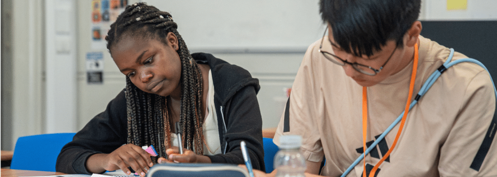 A picture of secondary pupils engaged in their work during an Action Tutoring session. The pupils are looking down at their workbooks on their desk and are appearing thoughtful while holding pens in their hands. 