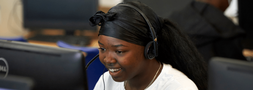 A picture of a pupil smiling and looking at a computer monitor during an Action Tutoring session. The pupil is wearing headphones while also wearing a headscarf and white t-shirt. 