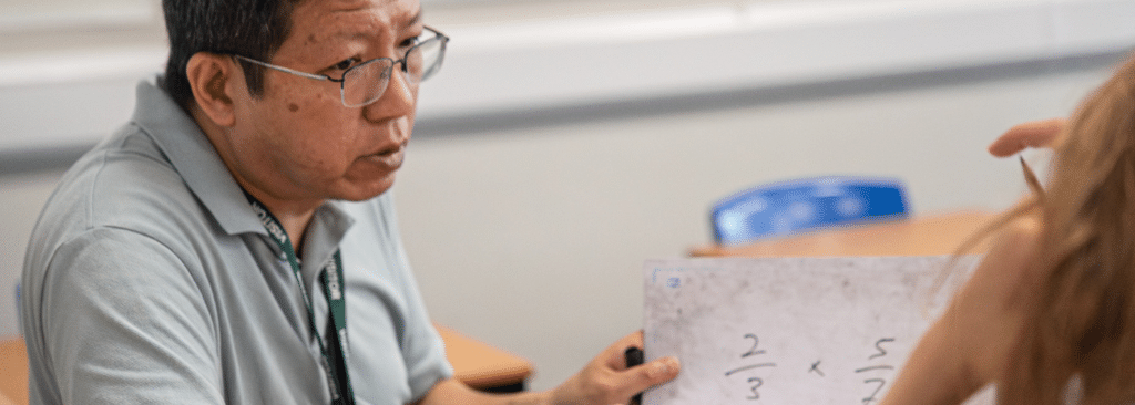 A picture of a tutor delivering a secondary maths session on an Action Tutoring programme. The tutor is holding a whiteboard which has some maths formulas on it and is looking thoughtfully across at the pupil. 