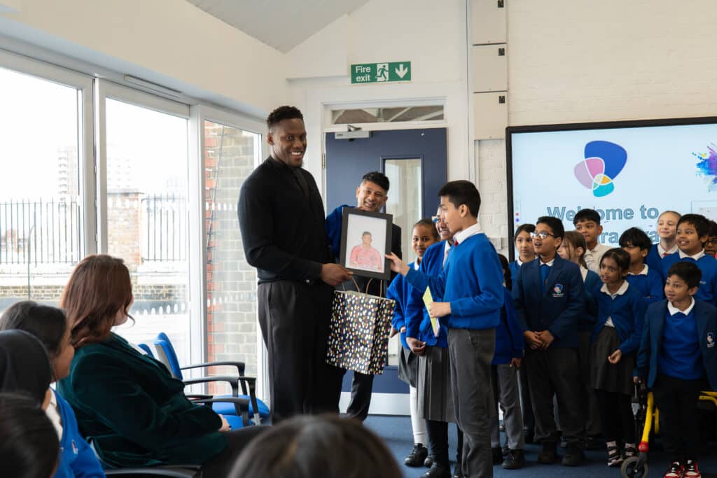 A primary school pupil giving Maro Itoje a drawing of him. The pupil's classmates are standing around them smiling.